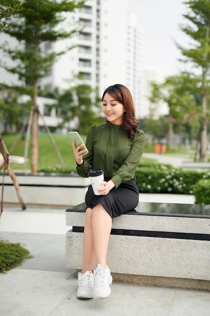 Beautiful business woman in casual green outfit sitting in park, holding coffee and talking on a phone