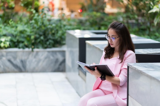 Beautiful business woman in glasses in a pink suit sits on a marble bench with a notebook and a pen in her hands something is writing down