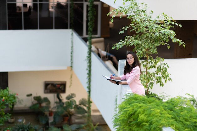 Beautiful business woman in glasses in a pink suit stands on a balcony surrounded by green plants with a notebook and a pen in her hands looks at the camera