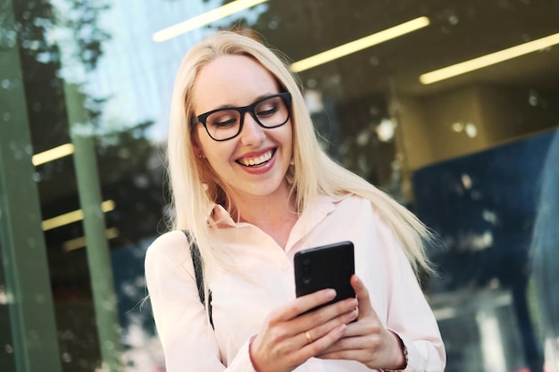 Beautiful business woman in a shirt black skirt and glasses at the panoramic window of the business center holds a mobile phone in her hands rejoices and smiles Free time spent on social networks