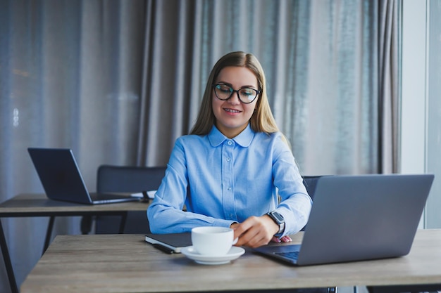 A beautiful business woman in a shirt and glasses sits with a laptop in the office at the table Woman manager in glasses at workplace in modern office Working day at the computer