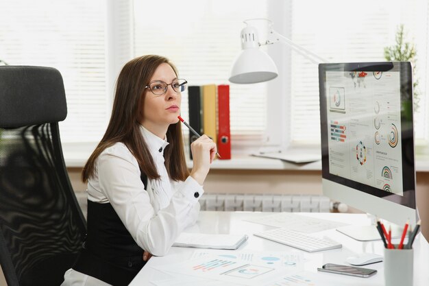 The beautiful business woman in suit and glasses working at computer with documents in light office, looking aside