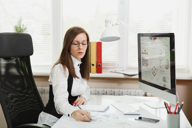 The beautiful business woman in suit and glasses working at computer with documents in light office, looking at the table