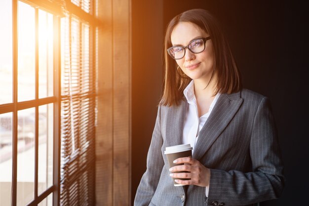 Beautiful business woman with coffee stands by the window, sunlight.