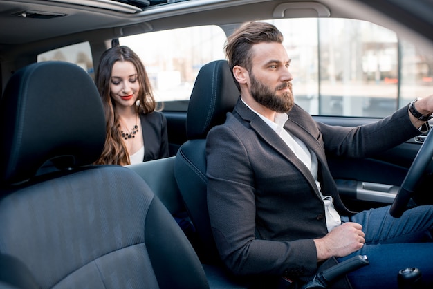 Beautiful businesswoman sitting on the backseat with elegant man driving the car in the city