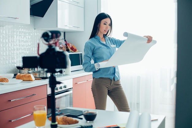 Beautiful calm woman standing in the kitchen and opening architectural paper roll Camera on tripod on the table