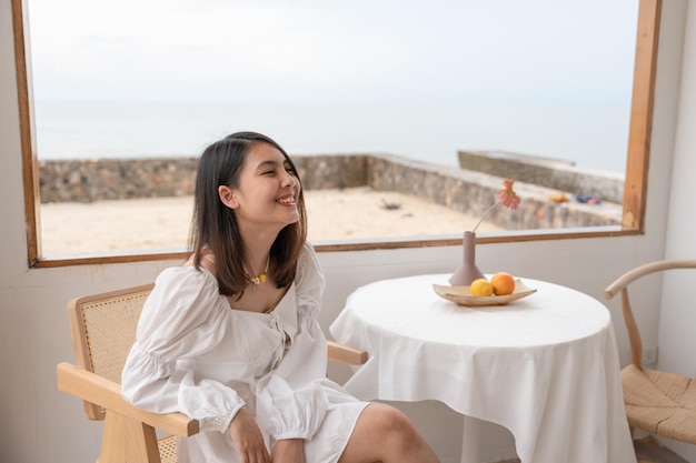 Beautiful carefree young asian woman in white dress sitting and smiling at table by window in the cafe on summer weekend