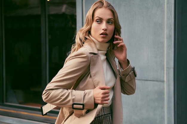 Beautiful casual blond girl in trench coat talking on cellphone thoughtfully looking away outdoor