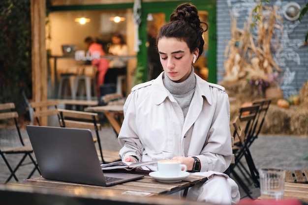 Beautiful casual brunette girl in trench coat with coffee studying on laptop in cafe on city street