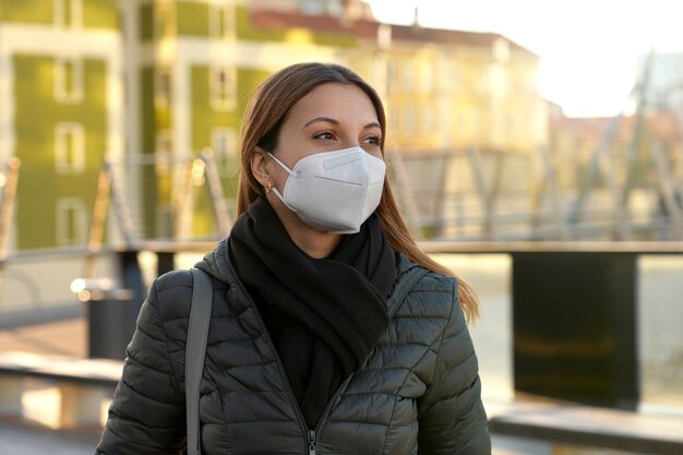 Beautiful casual young woman wearing a protective KN95 FFP2 mask walking in city street on sunset