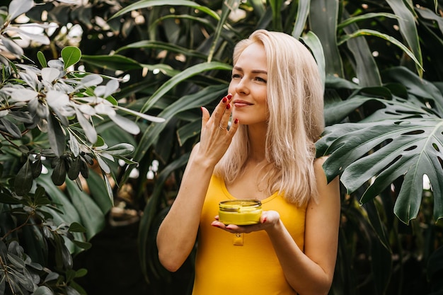 Beautiful caucasian blonde woman with healthy smooth clean skin holding jar of body cream and a happy contented face sniffs it against the background of exotic woods. cosmetic product concept