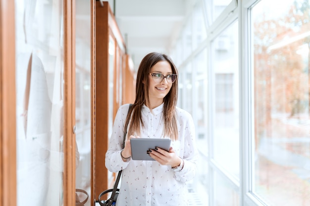 Beautiful Caucasian brunette next to noticeboard and using tablet while standing in university building and looking trough window..