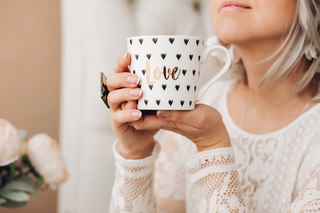 Beautiful caucasian female with short fair hair in white sweater holds a white cup of coffe or tea, drinks it and lies on the sofa