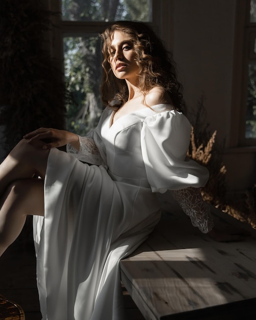 Beautiful caucasian girl bride with curly red hair sits in a wedding dress among dry flowers indoors on the table in a fashion pose and looks at the camera