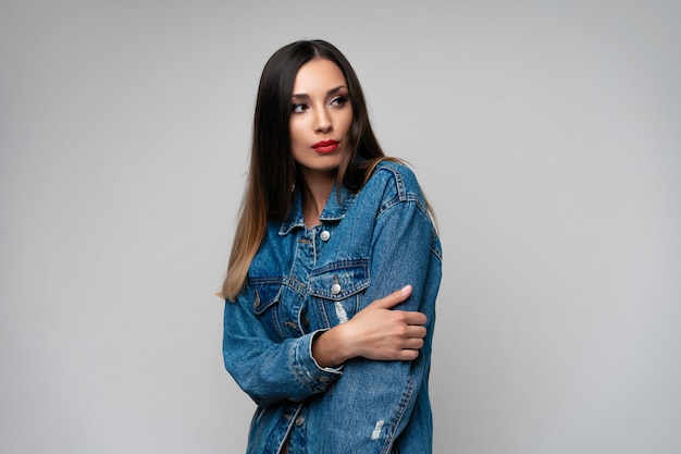 Beautiful caucasian girl denim jacket posing in studio on white background. Cute brunette. Denim fashion red lips elegant make up. Young adult woman standing indoors