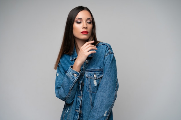 Beautiful caucasian girl denim jacket posing in studio on white background. Seductive brunette. Denim fashion red lips elegant make up. Young adult woman standing indors with sad face emotins