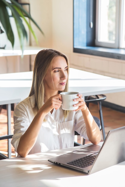 Beautiful caucasian girl drinks coffee and works on a laptop remotely in a bright space with green plants.