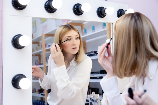 Beautiful caucasian girl make up artist, visagiste, model look at reflection in mirror with lamps and applying black lash mascara.  advert no brand decorative professional cosmetic store