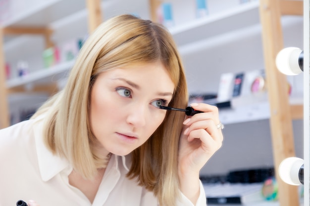 Beautiful caucasian girl make up artist, visagiste, model look at reflection in mirror with lamps and applying black lash mascara.  advert no brand decorative professional cosmetic store