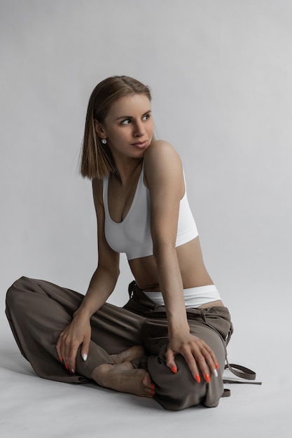 Beautiful caucasian girl in white lingerie and trousers sits on a white background in the studio and smiles