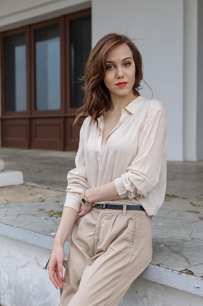 Beautiful caucasian girl with red lips and curly hair in stylish beige blouse looks at camera, posing outdoors. attractive female street portrait at spring. lifestyle, stylish fashion outfit