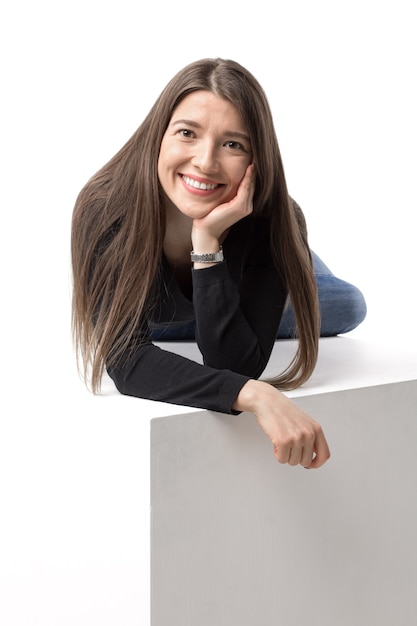 Beautiful caucasian lady is lying on the cube and smiling to a camera. Isolated image on the white wall.