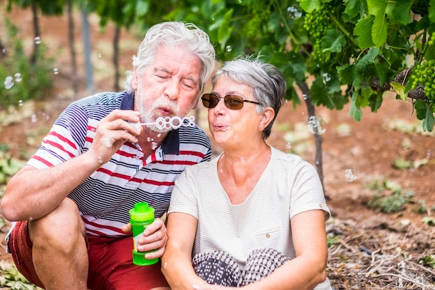 beautiful caucasian mature couple man and woman do soap bubbles together to play and have fun with joy, outdoor nature location for happy leisure activity for retired people with lifestyle