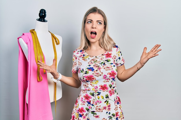 Beautiful caucasian woman dressmaker designer by manikin crazy and mad shouting and yelling with aggressive expression and arms raised. frustration concept.