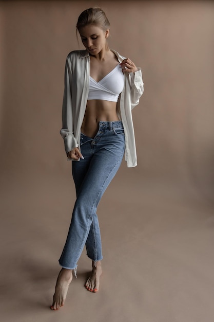 Beautiful caucasian woman in white lingerie shirt and jeans on a beige background walks and look down