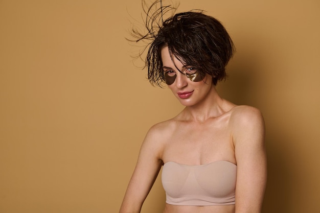 Beautiful Caucasian woman with healthy clean shiny skin, in beige underwear, wearing smoothing collagen under eyes patches and wet hair looking at camera, isolated on beige background with copy space