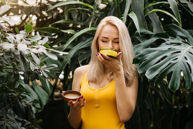 Beautiful caucasian woman with healthy smooth clean skin holding jar of body cream, sunblock, smelling its scent with closed eyes, posing in rainforest. Skincare, spa, natural cosmetic product concept