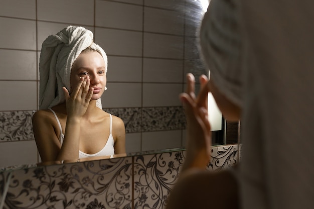 Beautiful Caucasian woman with a towel on her head in the bathroom applies cream after a shower to her face