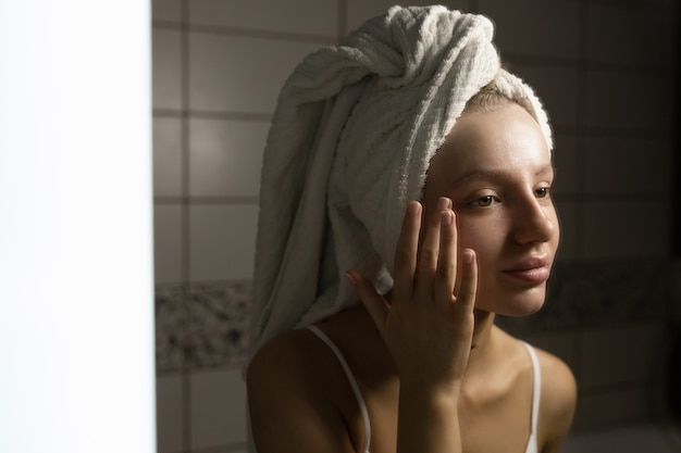 Beautiful caucasian woman with a towel on her head in the bathroom looks in the mirror and applies cream to her face