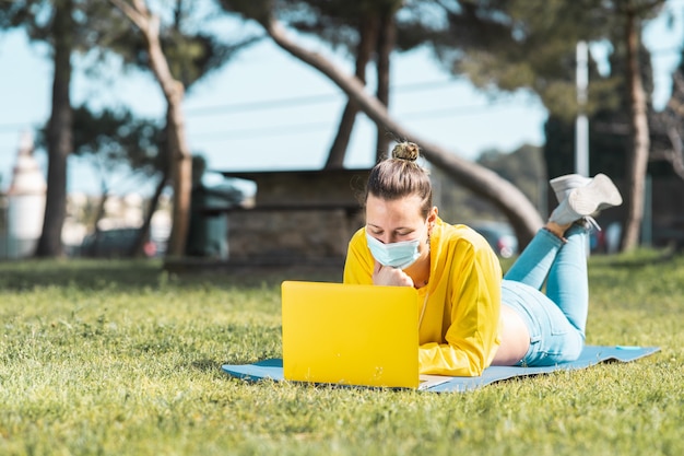 Beautiful Caucasian woman in a yellow crop jacket and face mask using a laptop in the park