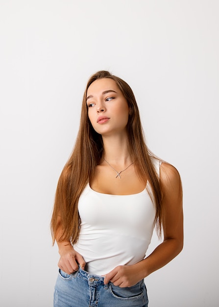 A beautiful caucasian young slender long haired brunette girl holds her jeans and looks thoughtfully to the side. The concept of healthy food, diet, lifestyle.  on white wall