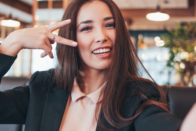 Beautiful charming brunette smiling asian girl taking selfie on frontal camera at cafe