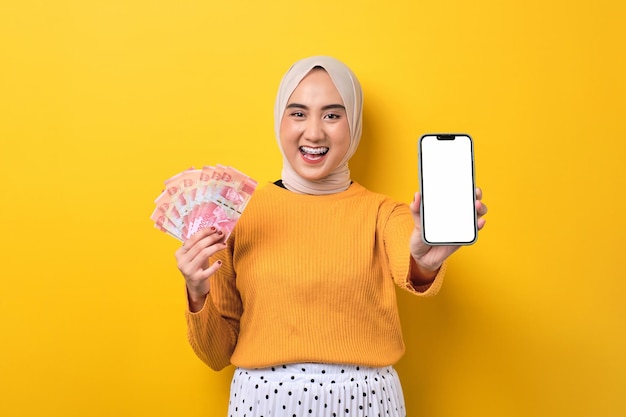 Beautiful cheerful Asian girl wearing hijab showing mobile phone with blank white screen holding cash money isolated on yellow background mockup copy space