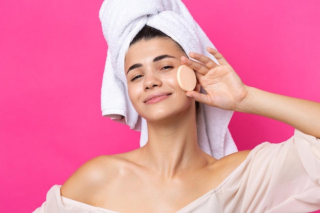 Beautiful cheerful attractive girl with a towel on her head holds a sponge against a pink background