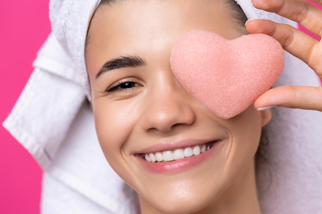 Beautiful cheerful attractive girl with a towel on her head holds a sponge in the form of a pink heart