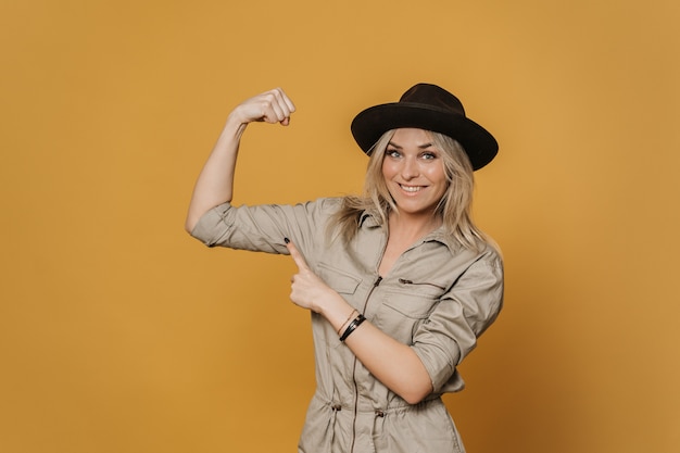 Beautiful cheerful  blonde woman in casual overalls and hat smiling widely, demonstrates her biceps and pointing on it by another hand, looks happily, against yellow backdrop. Positive people concept.