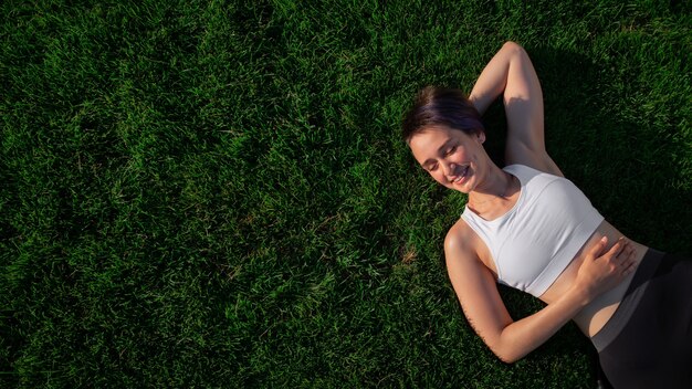 A beautiful, cheerful, cheerful woman in a white T-shirt is lying on the green lawn in the park and smiling. Happy woman relaxing on the grass during sunny summer day. Top view. large format banner