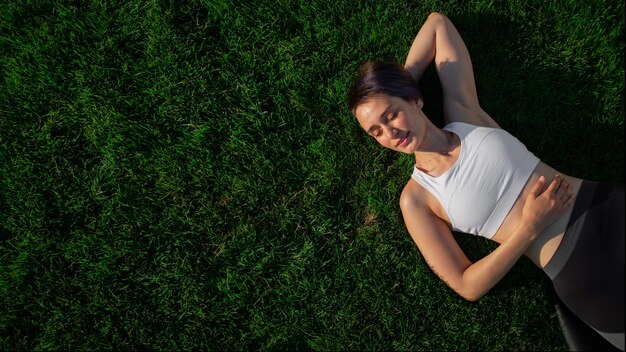 A beautiful, cheerful, cheerful woman in a white T-shirt is lying on the green lawn in the park and smiling. Happy woman relaxing on the grass during sunny summer day. Top view. large format banner