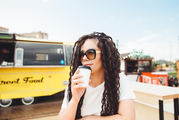 A beautiful cheerful girl with dreadlocks is sitting on a food court and drinking coffee fast food cafe on the street a woman drinks coffee in a cafe autumn