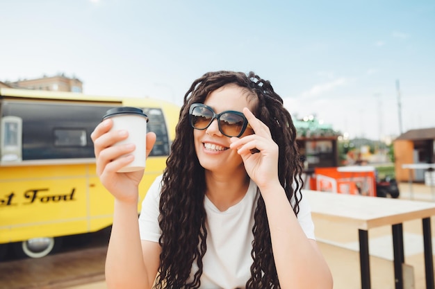 A beautiful cheerful girl with dreadlocks is sitting on a food court and drinking coffee fast food cafe on the street a woman drinks coffee in a cafe autumn