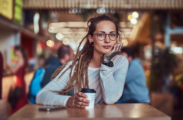 Beautiful cheerful girl with tattooes and dreadlocks is sitting at food court while drinking coffee.