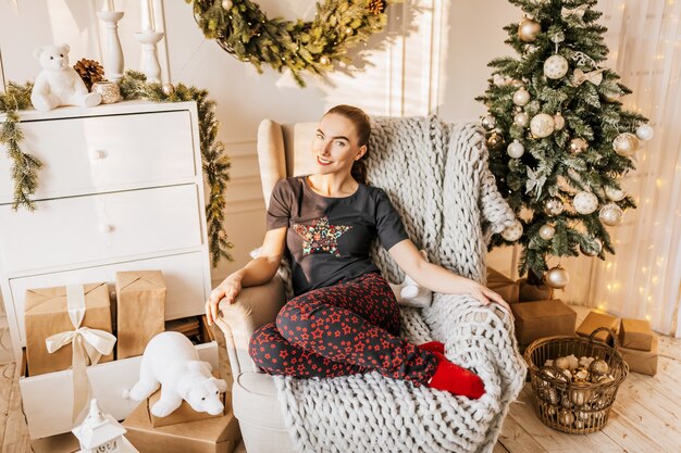 Beautiful cheerful happy young girl in pajamas with christmas gifts on sofa on the of a new year tree at home