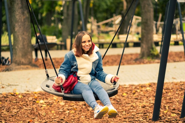 A beautiful and cheerful woman in warm clothes swings on a swing in the park Girl swinging in the autumn city park