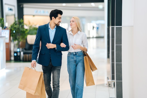 Beautiful cheerful young couple holding shopping paper bags with purchases and walking holding hands at mall. Handsome bearded man and attractive blonde woman purchasing together at store center.