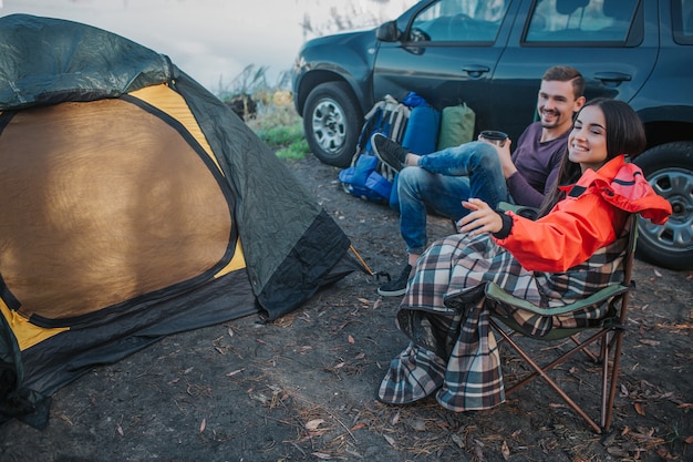 Beautiful and cheerful young woman points on left. They looks with guy in the same direction. People hold thermocups in hands. They sits on folding chairs at tent and car.