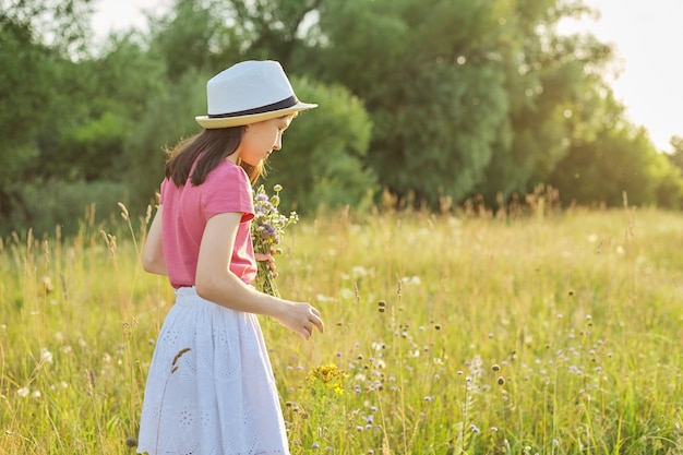 Beautiful child girl in hat, dress in field tearing bouquet of wildflowers, golden hour, with copy space. Beauty, nature, leisure, happy childhood, summer vacation concept